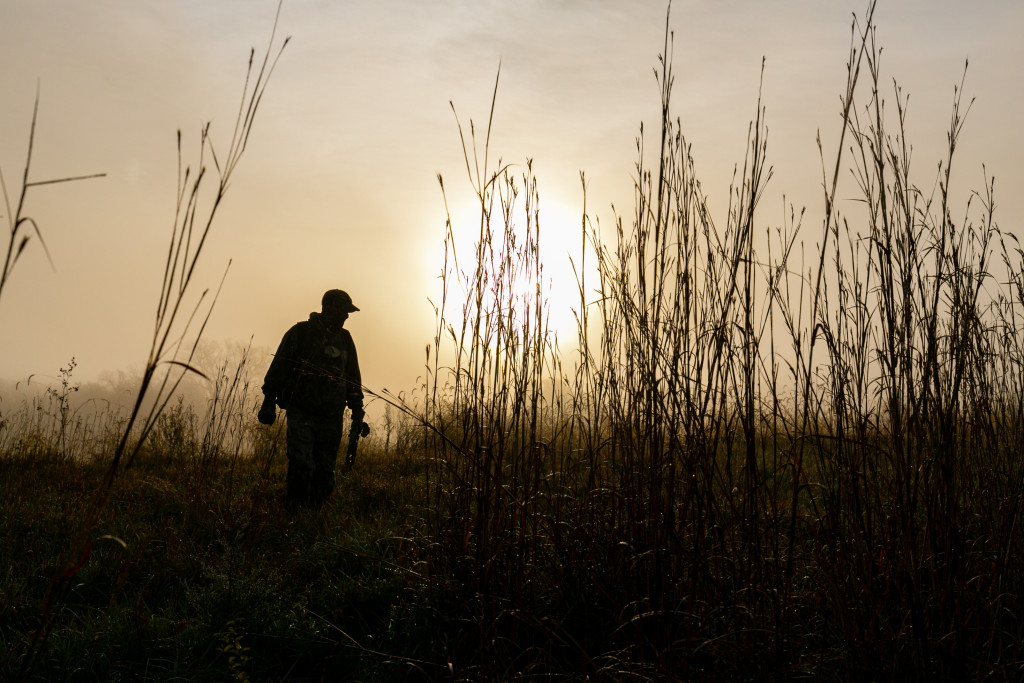 a man is silhouetted against a low, hazy sun walking along a tall-grass prairie