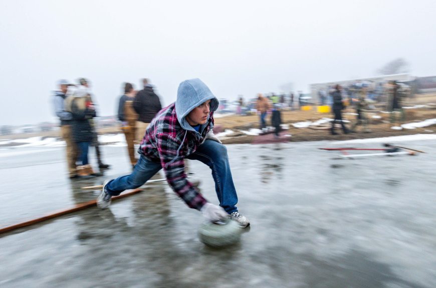 a man, his foot against a board to stop him slipping, pushes a curling stone across the ice