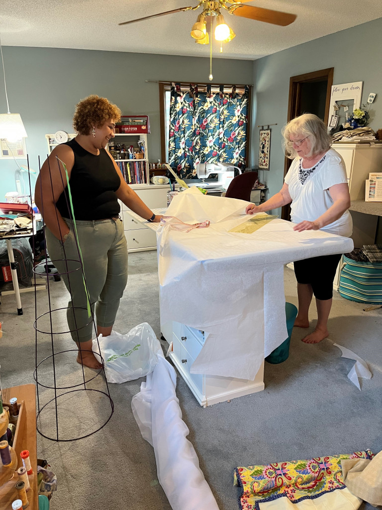 two women stand in a fabric shop cutting thin white gauze to cover an inverted tomato cage