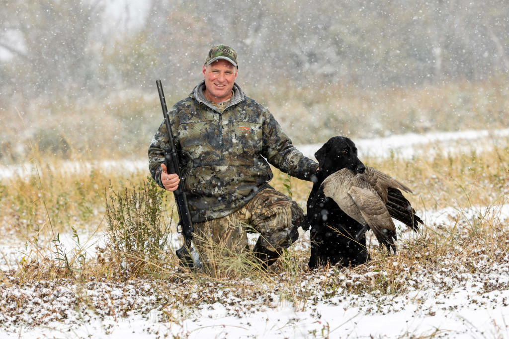 A male hunter kneels on the ground with a gun, his lab and goose harvest