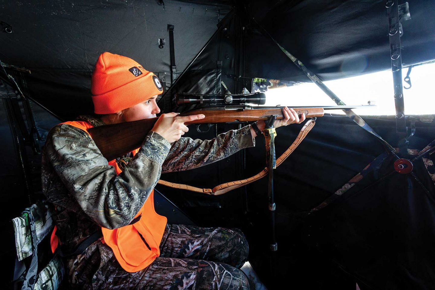 A young woman looking through the sight of rifle while deer hunting inside a ground blind.