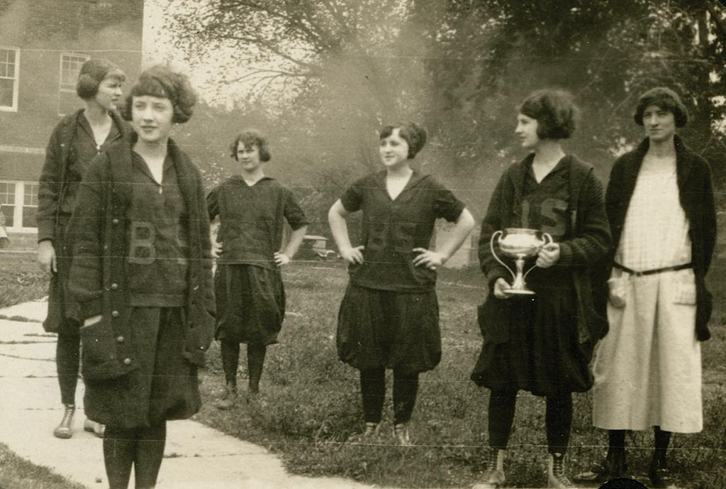 a black and white photo of girls in black dresses and athletic shoes holding a large silver trophy