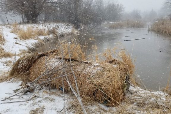 A bale of hay along a snowy river bank during winter as snow falls.