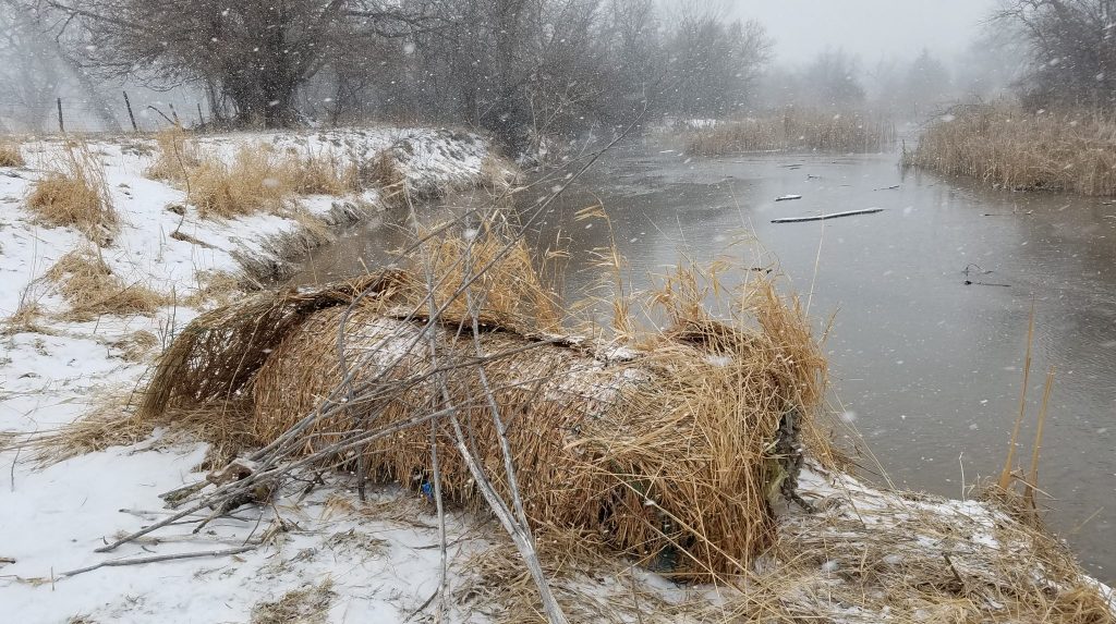 A bale of hay along a snowy river bank during winter as snow falls.