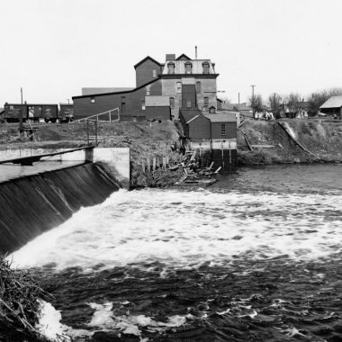 Water pours over a dam, a flour mill in the background