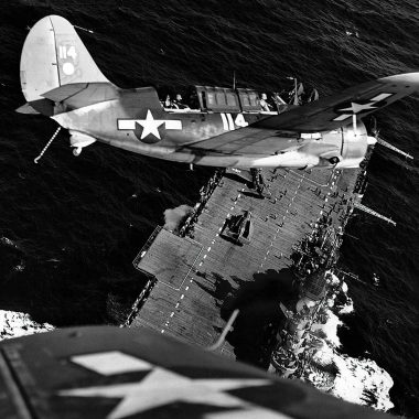 A black and white photo captures a Helldiver airplane turn its nose down to an aircraft carrier below floating on a calm ocean