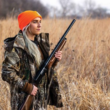 A blond woman in an neon orange stocking cap wears camo and walks through a tallgrass prairie