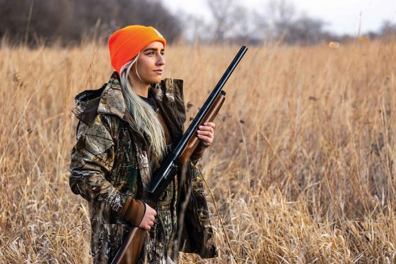 A blond woman in an neon orange stocking cap wears camo and walks through a tallgrass prairie