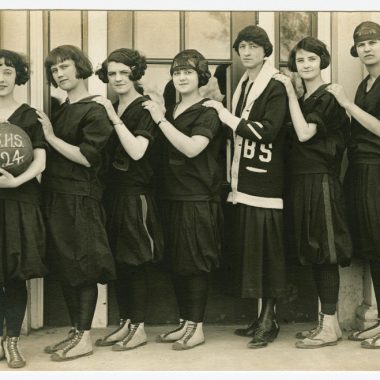 Seven girls stand in a row holding a basketballball with B.S.H.S. 1924 written on it. They wear black tights and balloon-bottom dresses.