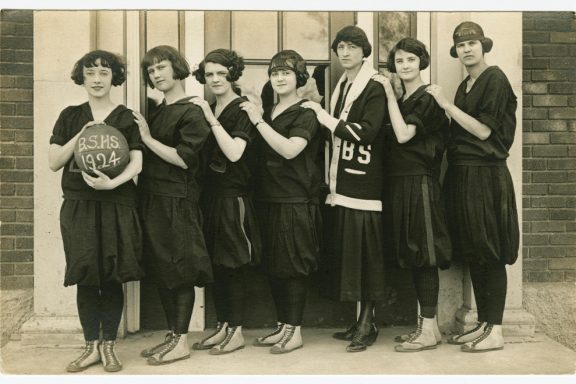 Seven girls stand in a row holding a basketballball with B.S.H.S. 1924 written on it. They wear black tights and balloon-bottom dresses.