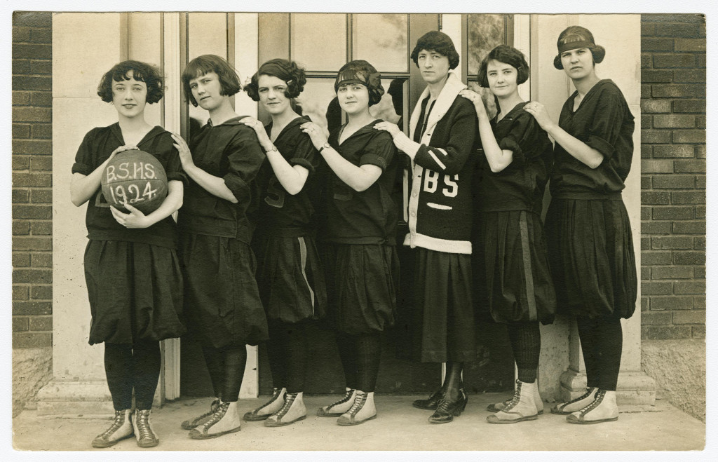 Seven girls stand in a row holding a basketballball with B.S.H.S. 1924 written on it. They wear black tights and balloon-bottom dresses.