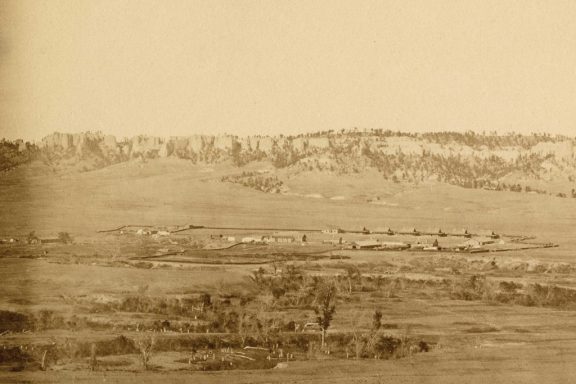 A historical sepia-toned photograph of tents set up small against a backdrop of buttes