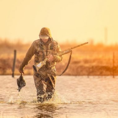 A man wades through water carrying a duck he shot while waterfowl hunting