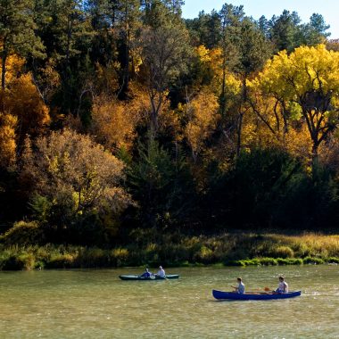 People canoeing and kayaking on a rive with autumn trees in the background.