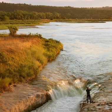 A man photographs the Niobrara river from water level near a small falls or beautiful orange light cascades across the scene. The viewpoint is from atop a hill looking down at this man photographing.