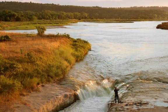 A man photographs the Niobrara river from water level near a small falls or beautiful orange light cascades across the scene. The viewpoint is from atop a hill looking down at this man photographing.