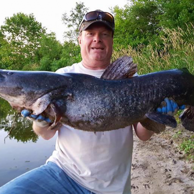 A man holds up a large flathead catfish at the edge of a sandpit lake.