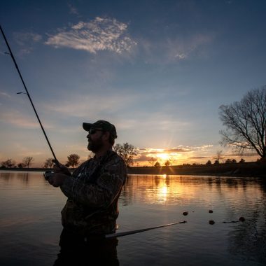 A man fishes in a lake wearing waders as the sun sets.