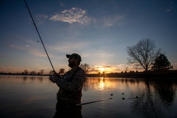 A man fishes in a lake wearing waders as the sun sets.