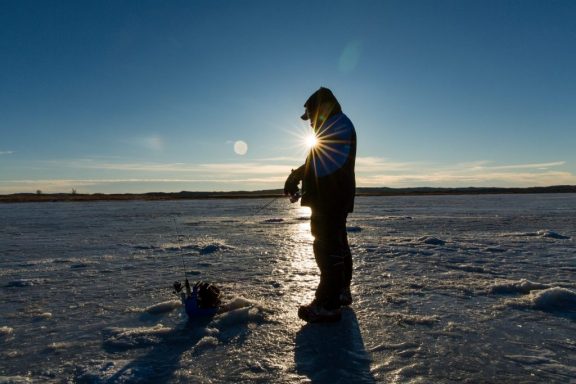 A man stands on a frozen lake preparing to ice fish with the sun beaming behind him.