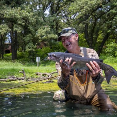 A man wearing waders in a lake smiles while holding a catfish he caught.