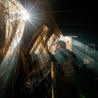 A combination of early-morning sun and smoke from breakfast in the goose blind make for interesting rays of light as a hunter peers out a duck blind.