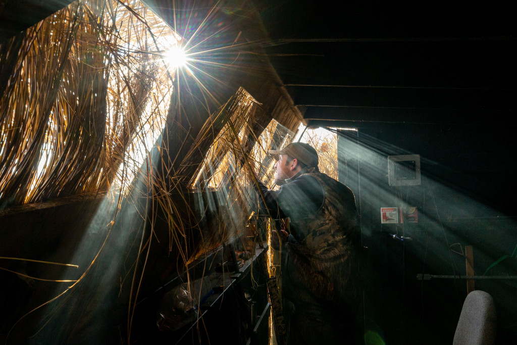 A combination of early-morning sun and smoke from breakfast in the goose blind make for interesting rays of light as a hunter peers out a duck blind.