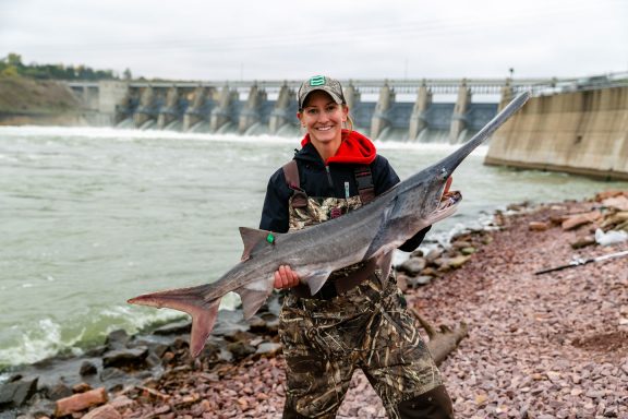 A woman smiles and holds a large paddlefish she caught near Gavins Point Dam.