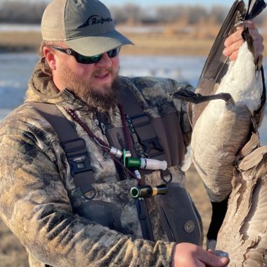 A man wearing duck calls around his neck holds a goose he harvested