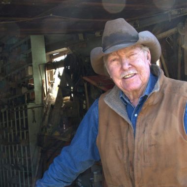 Wearing a blue, long-sleeved shirt, brown vest and beige cowboy hat, Paul Allen stands in the doorway of his barn-looking shop.