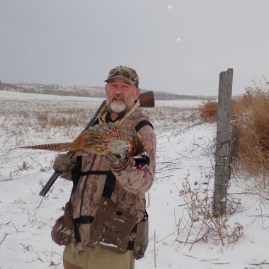 Daryl Bauer holding a pheasant while hunting in snow.