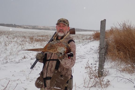 Daryl Bauer holding a pheasant while hunting in snow.