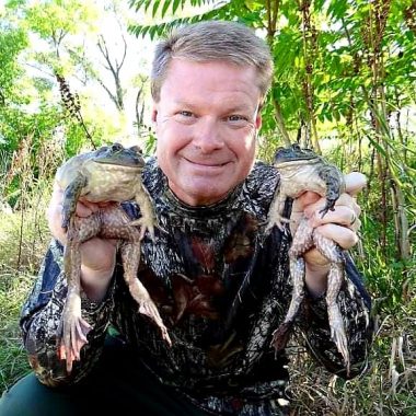 I man wearing camo holds two large American bullfrogs outdoors.