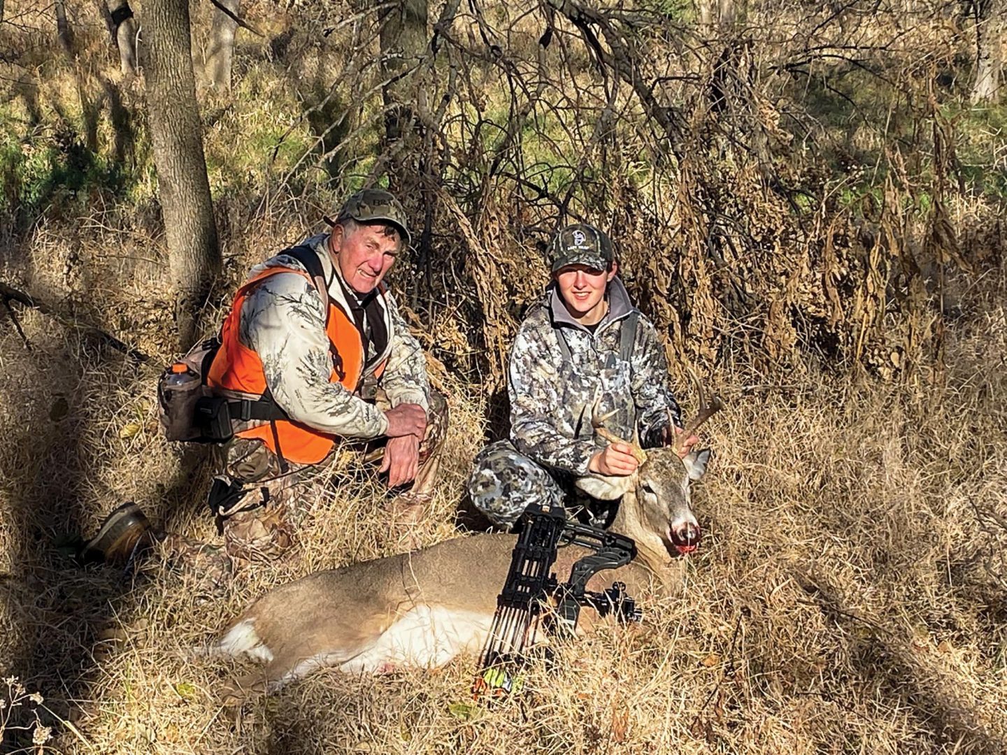 An older male hunter education instructor poses with a young woman and her deer on an archery hunt.