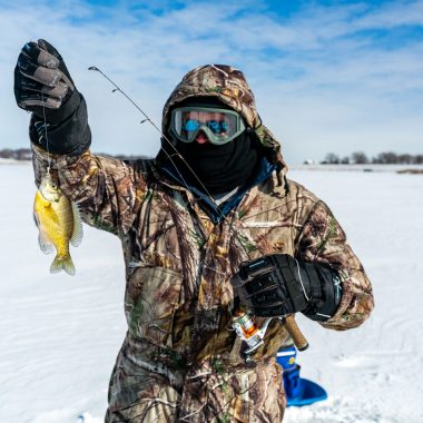 An angler in ice fishing gear, including snow goggles holding a bluegill on a frozen lake.