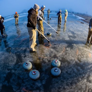 fish-eye lens used to capture scene on ice with people and their curling stones and brooms