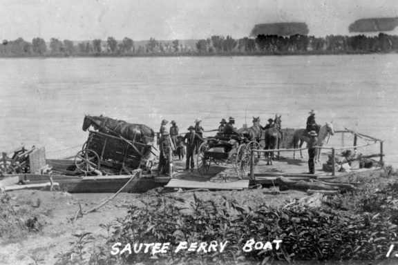 a rickety plank ferry with paddlewheels is loaded with wagons and carts